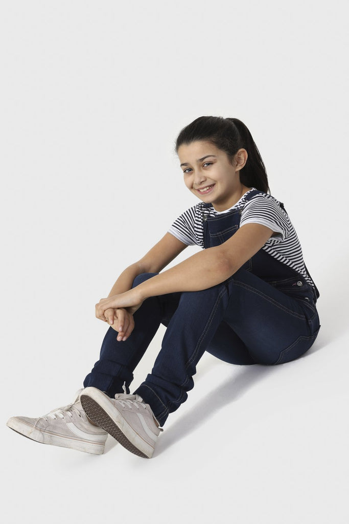 Girl sitting on floor with stretched legs, wearing slim fit denim dungarees with adjustable straps.