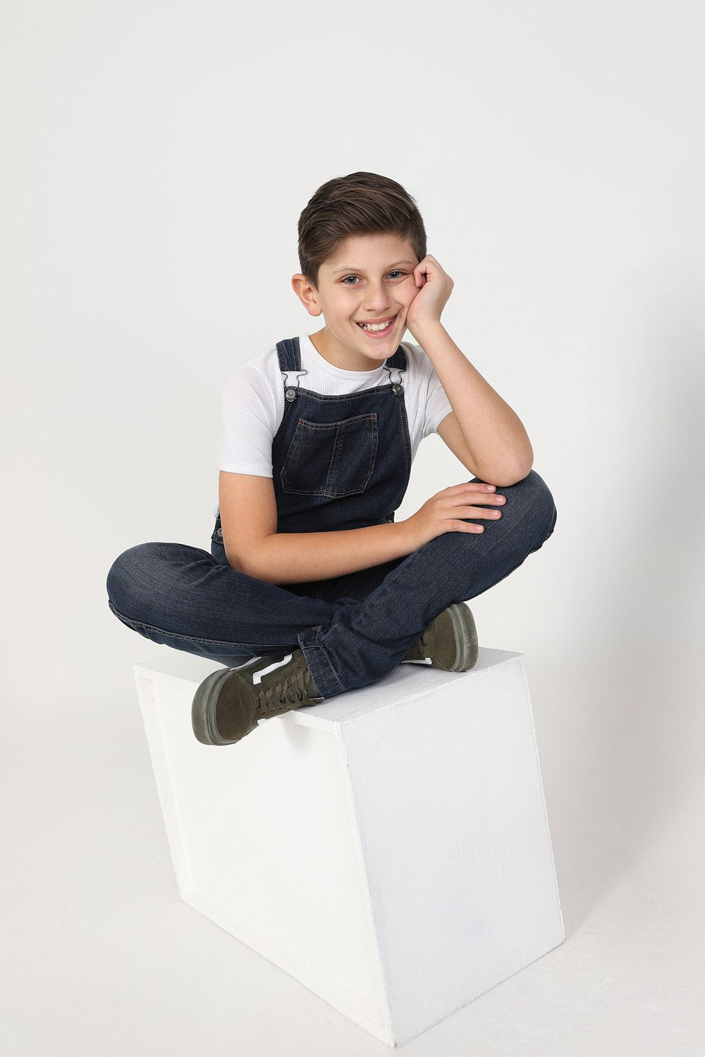 Smiling boy, cross-legged on box wearing modern, machine washable denim overalls.