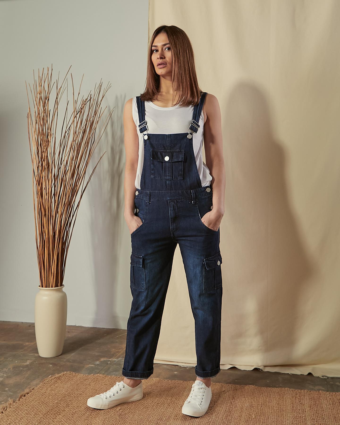 Full-length, front view of model wearing Tammy darkwash dungarees with hands in front pockets and view of large bib pocket.