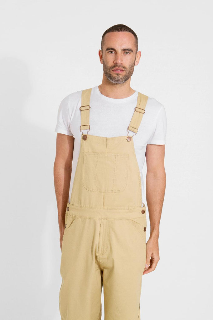 Frontal pose cropping bottom third, wearing loose fitting cotton dungaree shorts, with focus on side buttons and adjustable straps.
