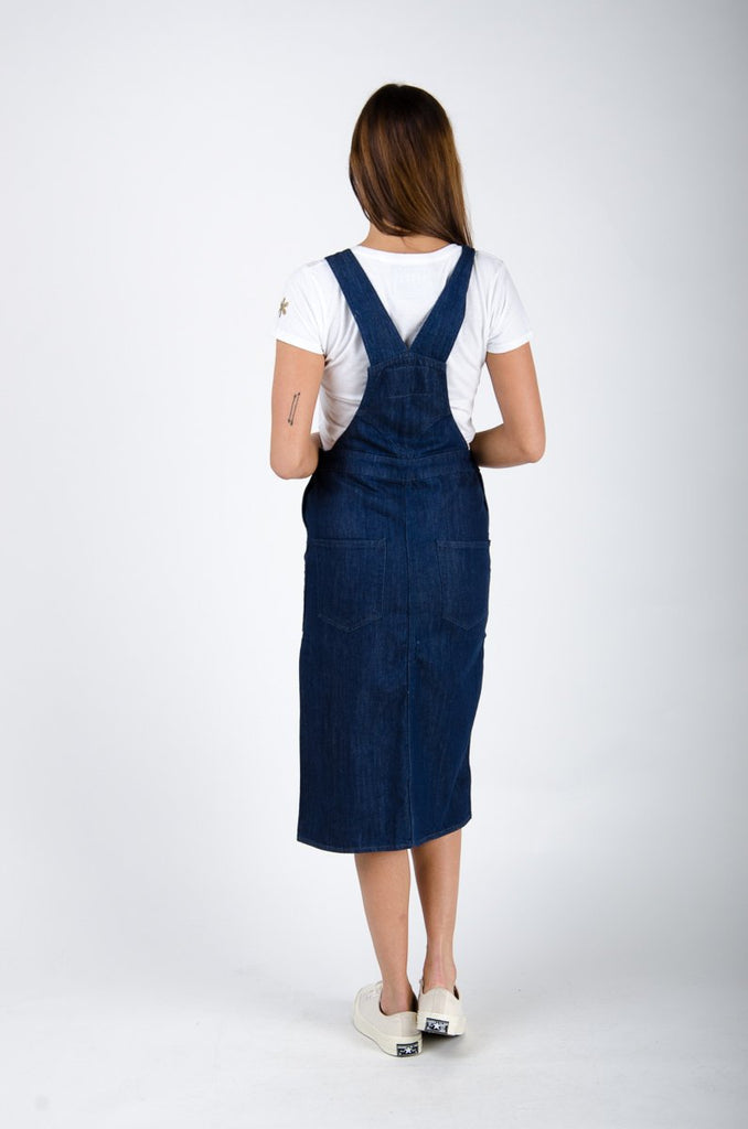 Full-length back shot of dark blue rinsed organic cotton bib-overall midi dress with view of back pockets and straps.