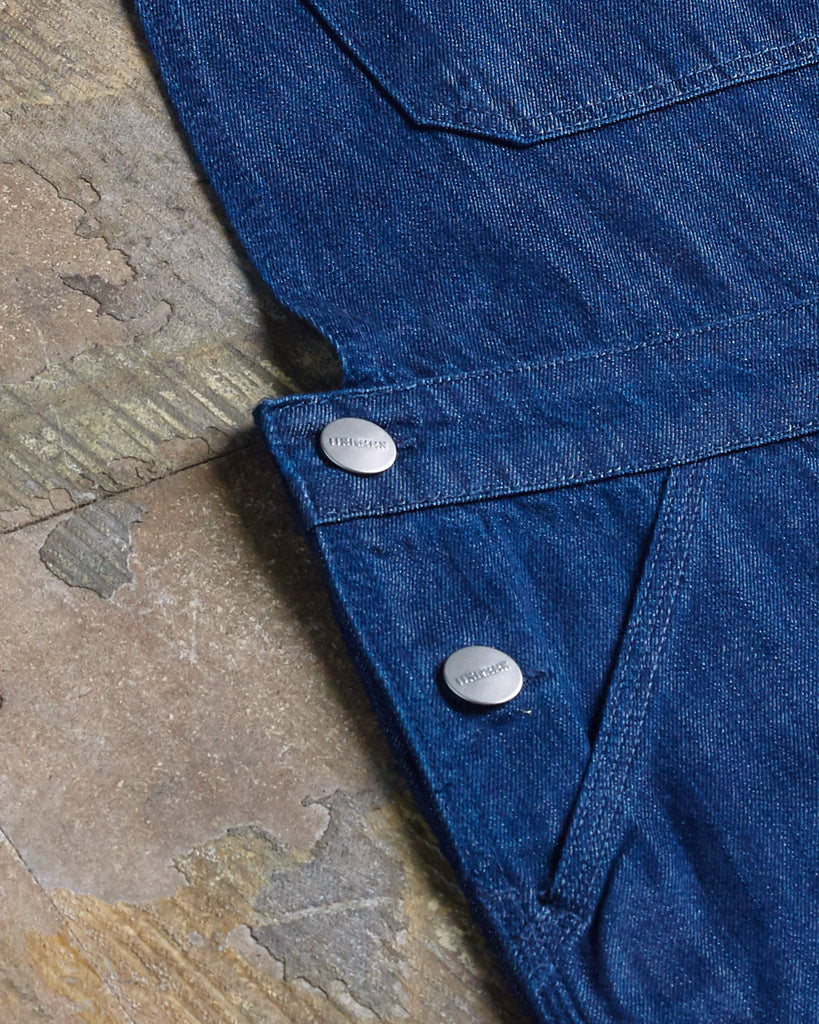 Close-up angled flat detail shot of button fastening and side pockets on dark blue rinsed denim midi dungaree dress.