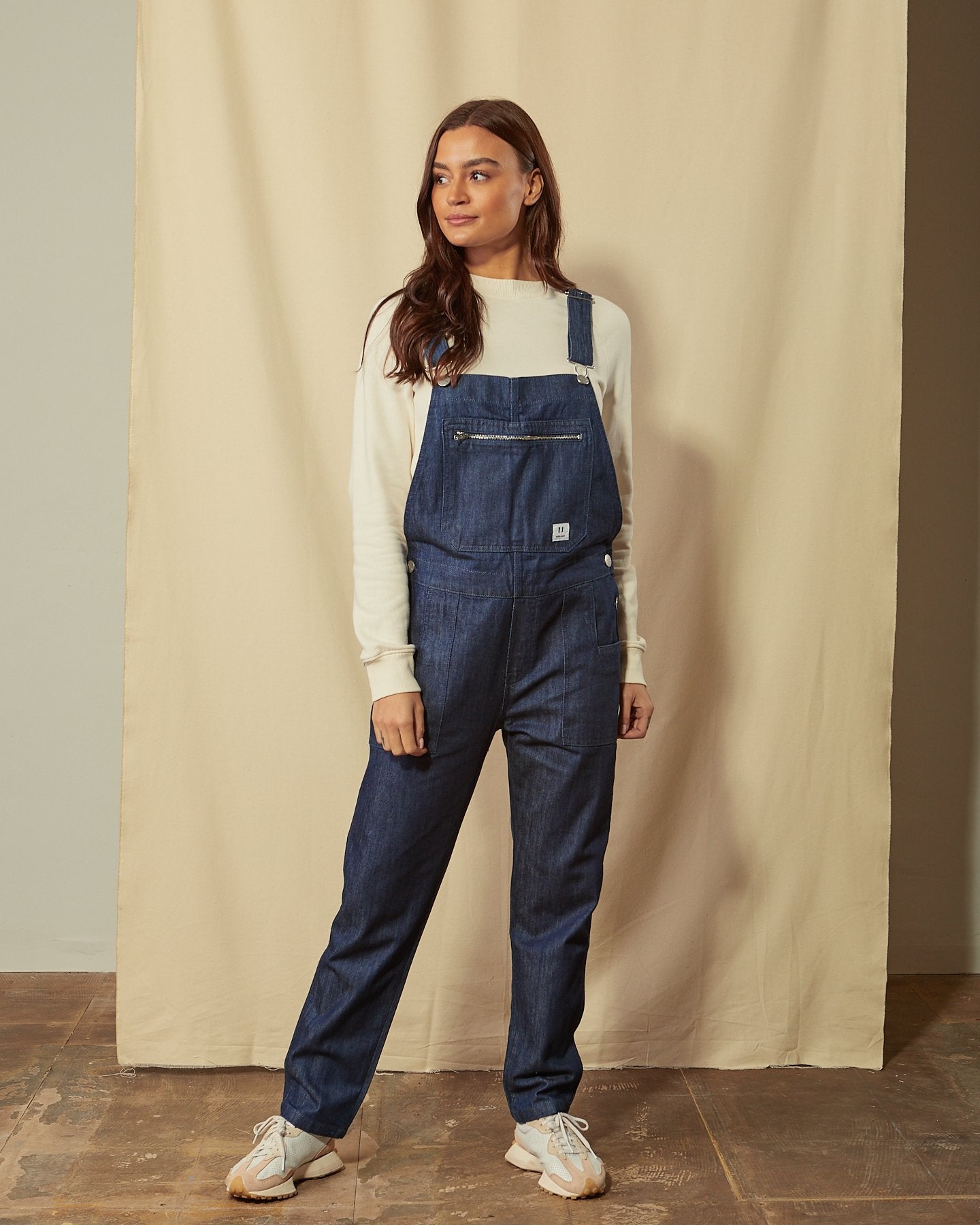 Front shot of women's dark blue rinsed organic denim bib-overalls with view of button closure, multiple pockets and adjustable straps. Model has hand in side pockets.