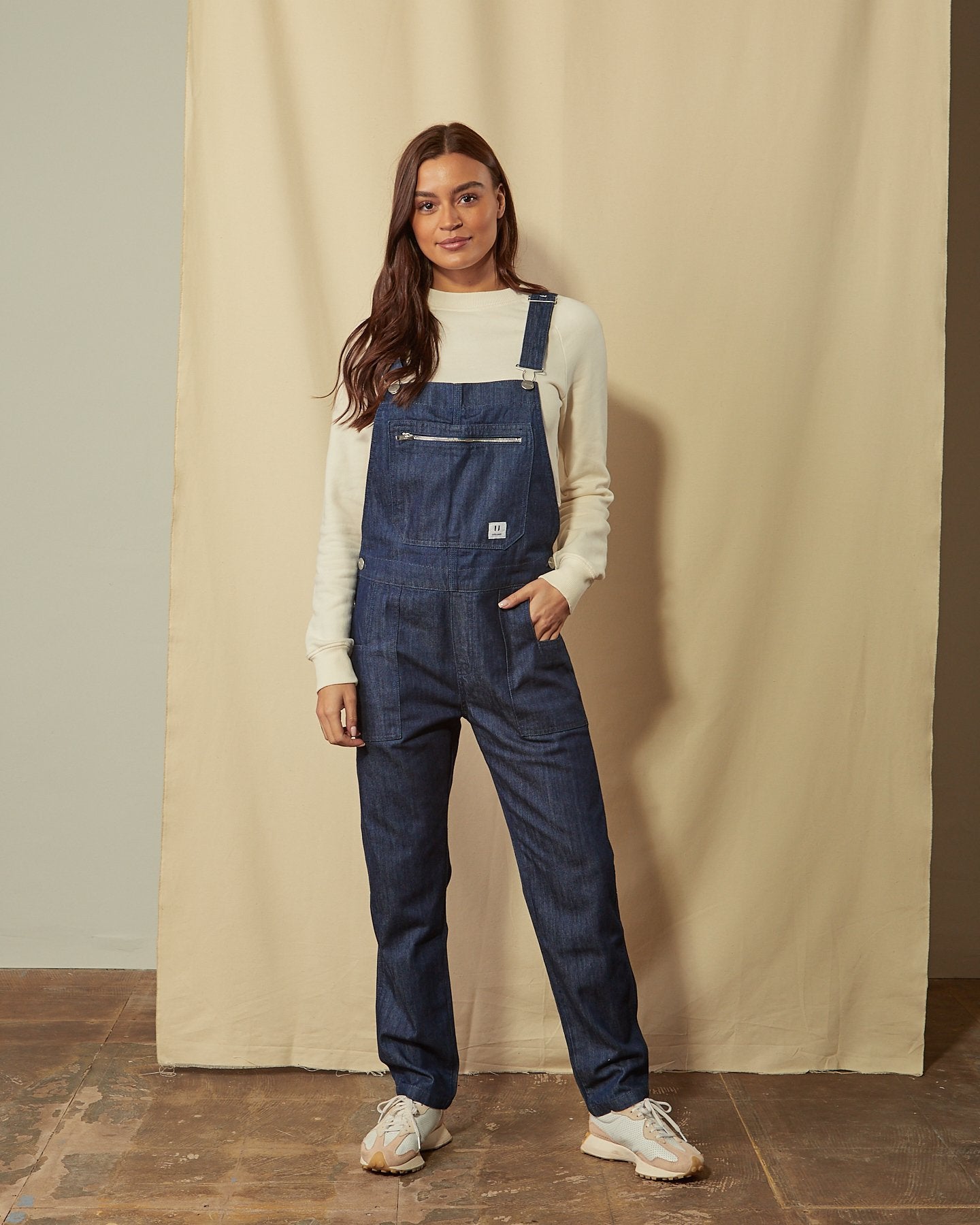 Full front view of model holding adjustable straps of Uskees men’s washed organic cotton utility dungarees with view of multiple pockets and button closure.