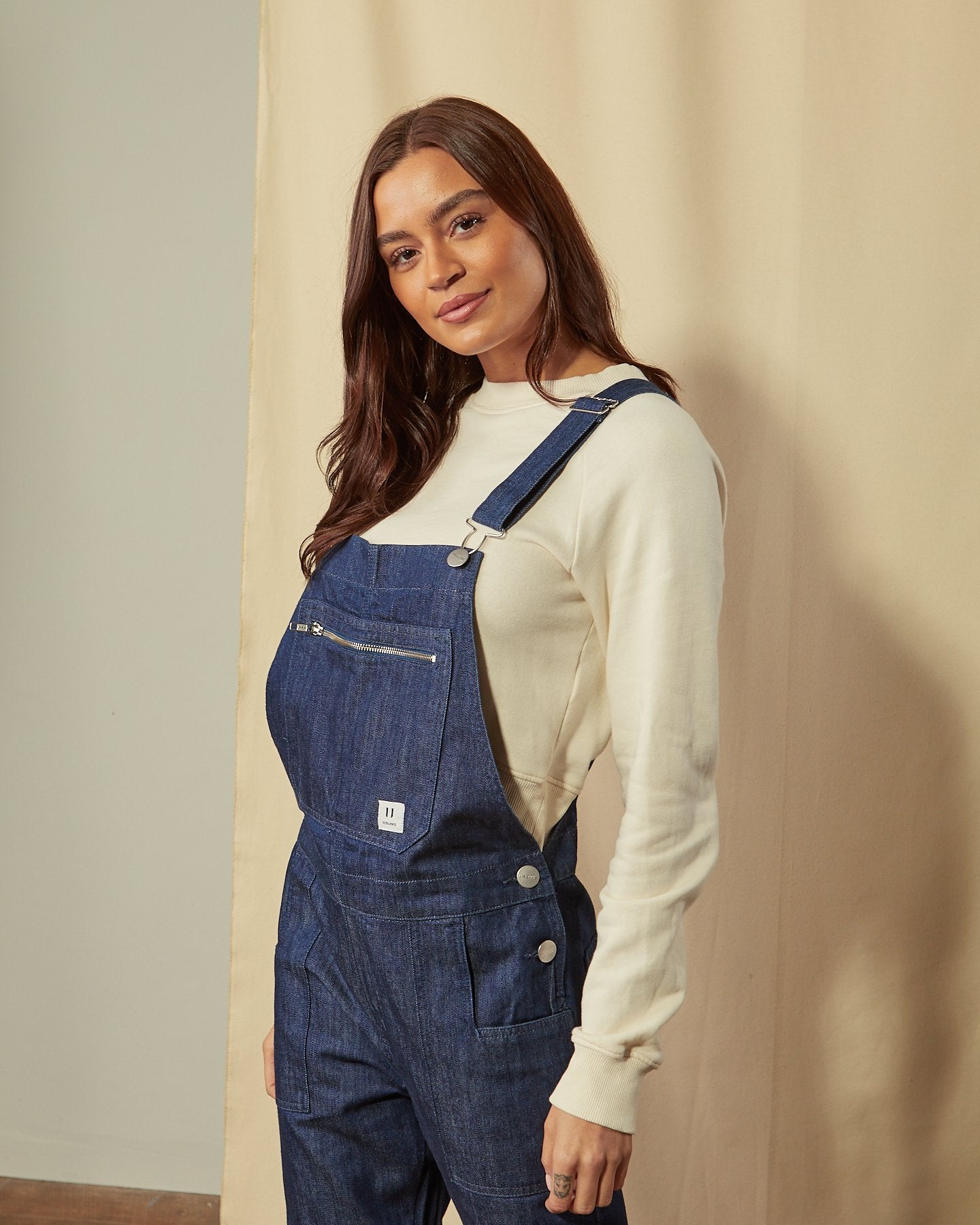 Full-length rear view of model wearing Uskees men’s washed denim utility bib-and-brace overalls with view of back pockets.