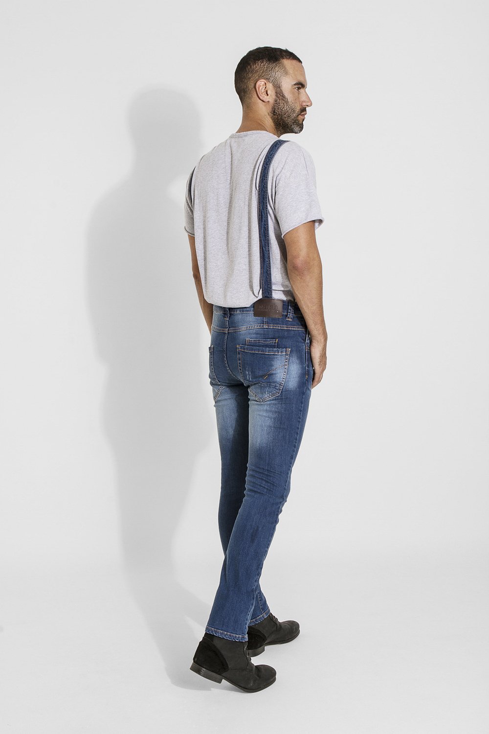 Full-length angled back walking pose of model wearing Whitefield grunge style blue denim denim bib overalls with view of back straps, back pockets and branding label.