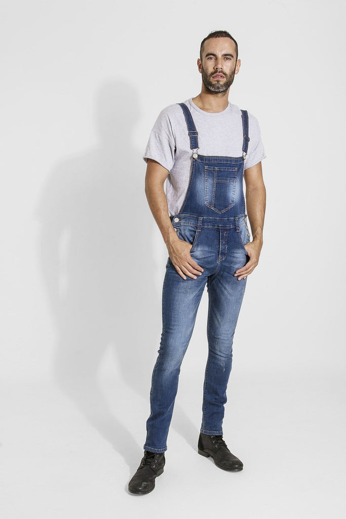 Full-length front view of model wearing Whitefield distressed style blue denim denim bib overalls with hands in front pockets.