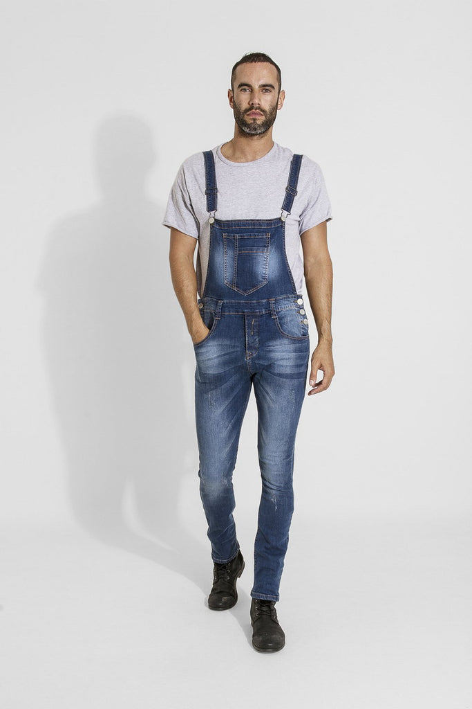 Full-length front walking pose of model wearing Whitefield distressed style blue denim denim bib overalls with hand in front pocket.
