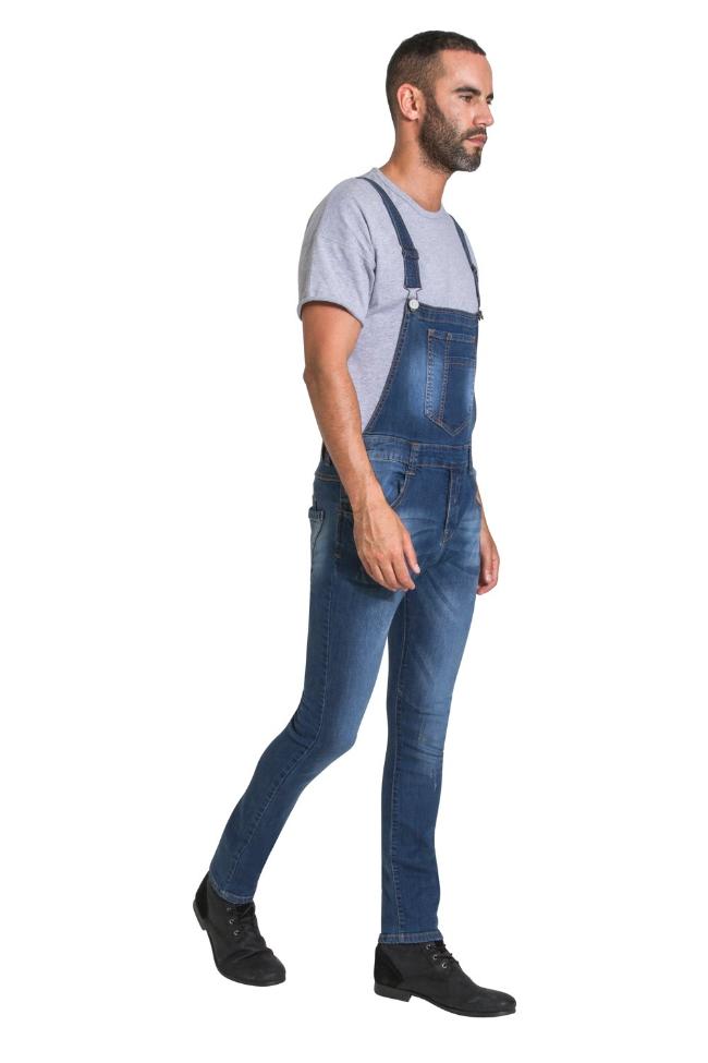 Full-length side walking pose of model wearing Whitefield distressed style blue denim denim bib overalls with view of multiple pockets.