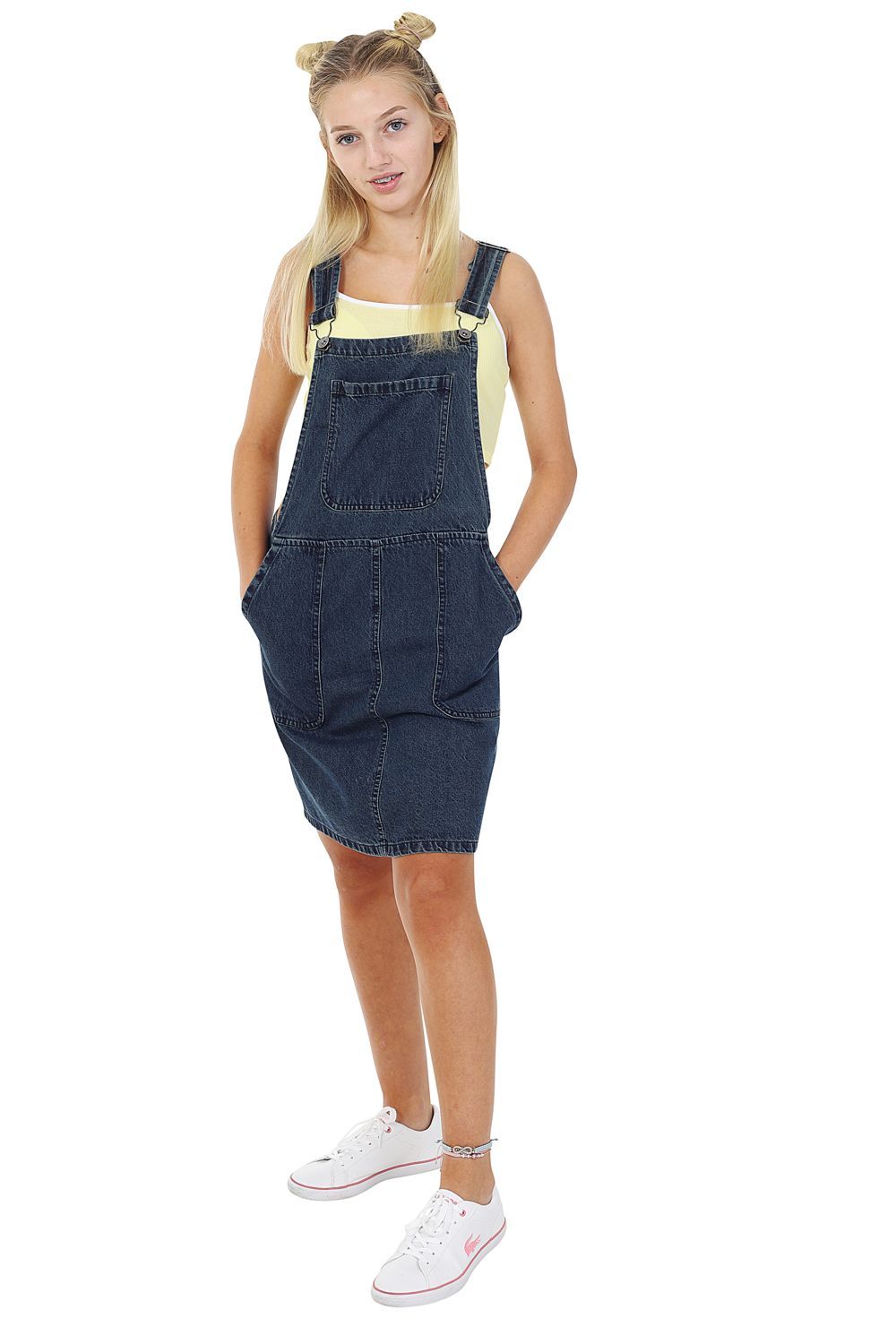 Full-length front view of model with hands in large front pockets of Winnie short denim dungaree dress in vintage wash, with view of bib pocket and adjustable straps.