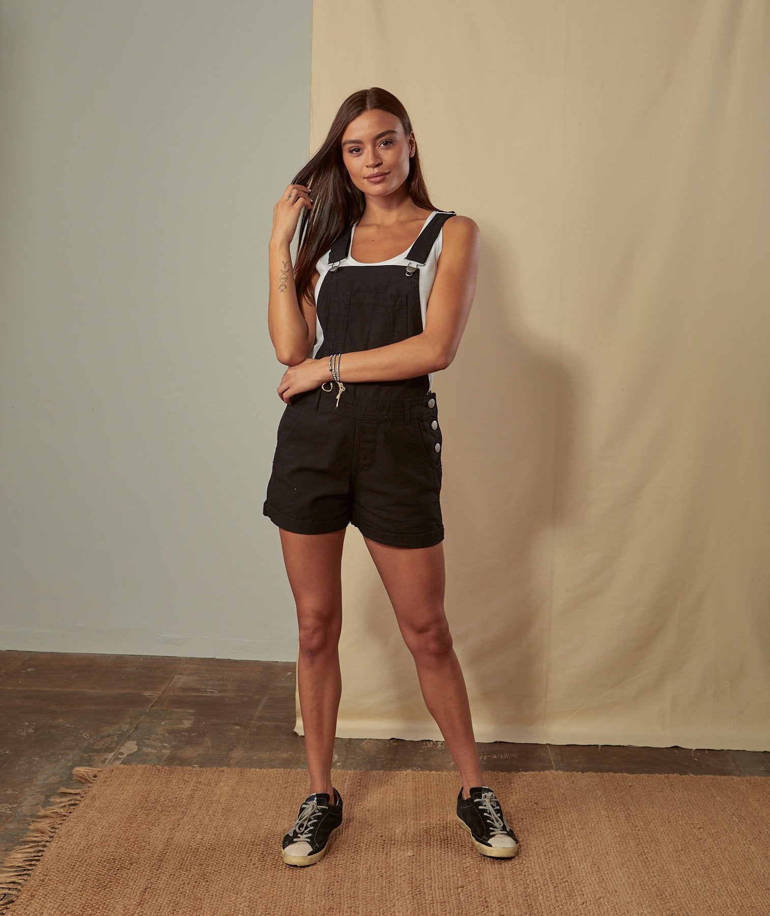 Full-length front view of model wearing Xenya black organic cotton dungaree shorts showing loose-fit silhouette and side-button fastening.