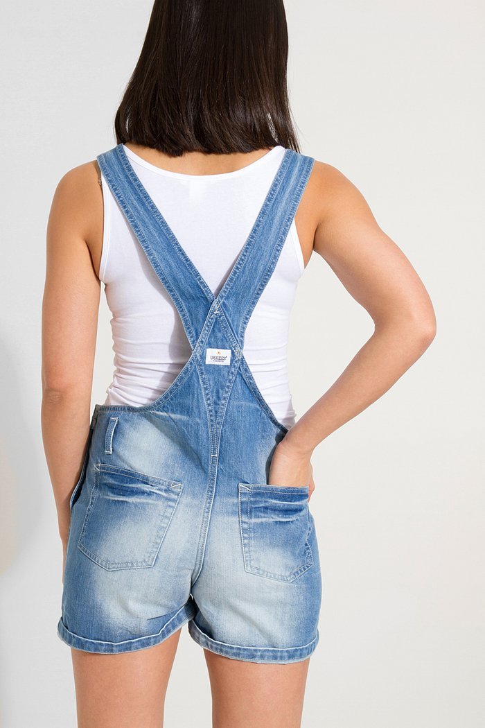 Two-thirds length back view of model wearing Xenya lightwash denim dungaree shorts showing back pockets and straps.