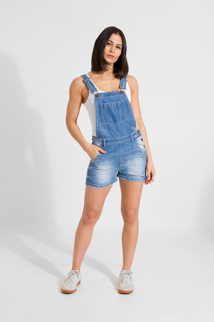 Full-length front view of model wearing Xenya lightwash denim dungaree shorts showing loose-fit silhouette and side-button fastening.