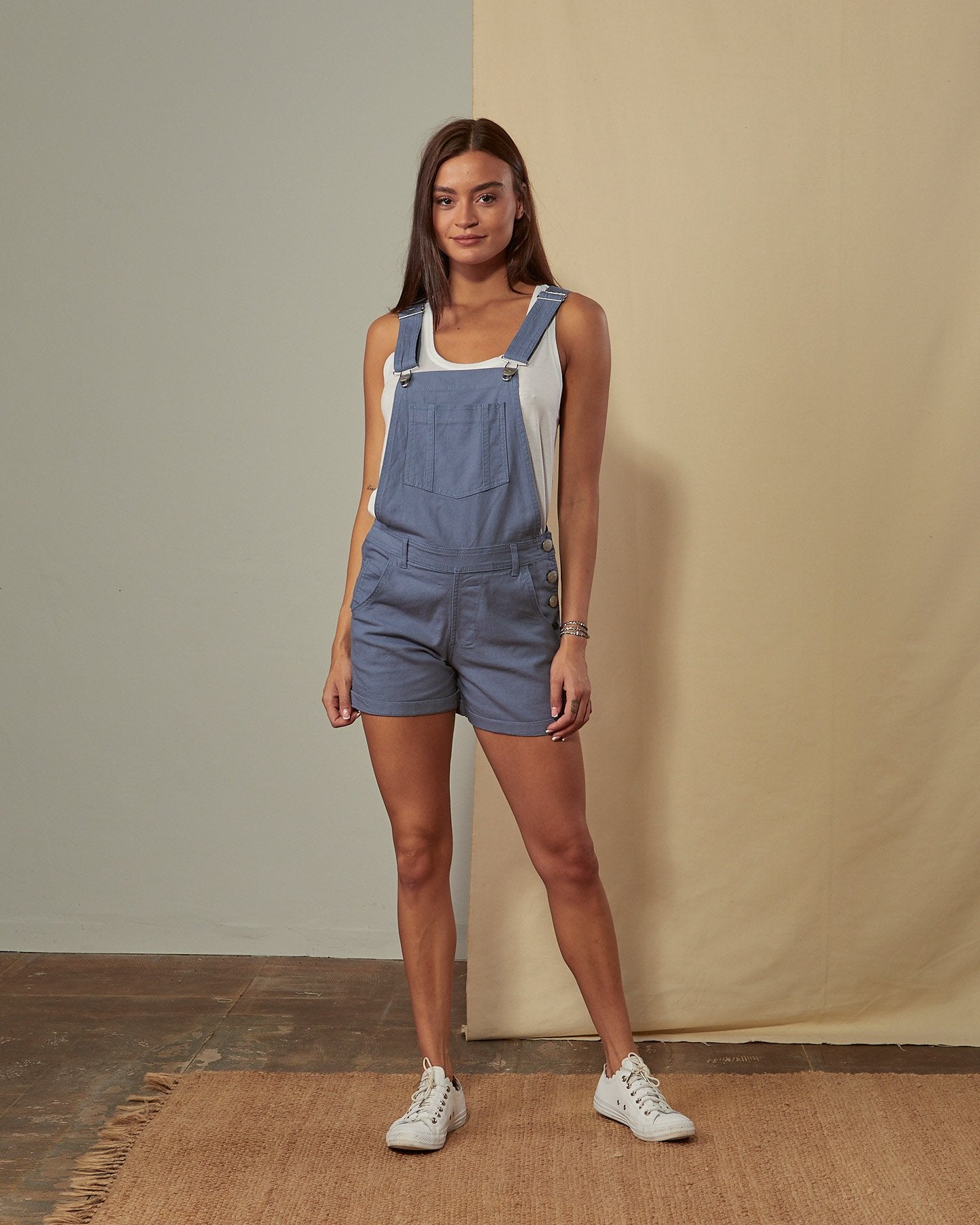 Full-length front view of model wearing Xenya teal organic cotton dungaree shorts showing loose-fit silhouette and side-button fastening.